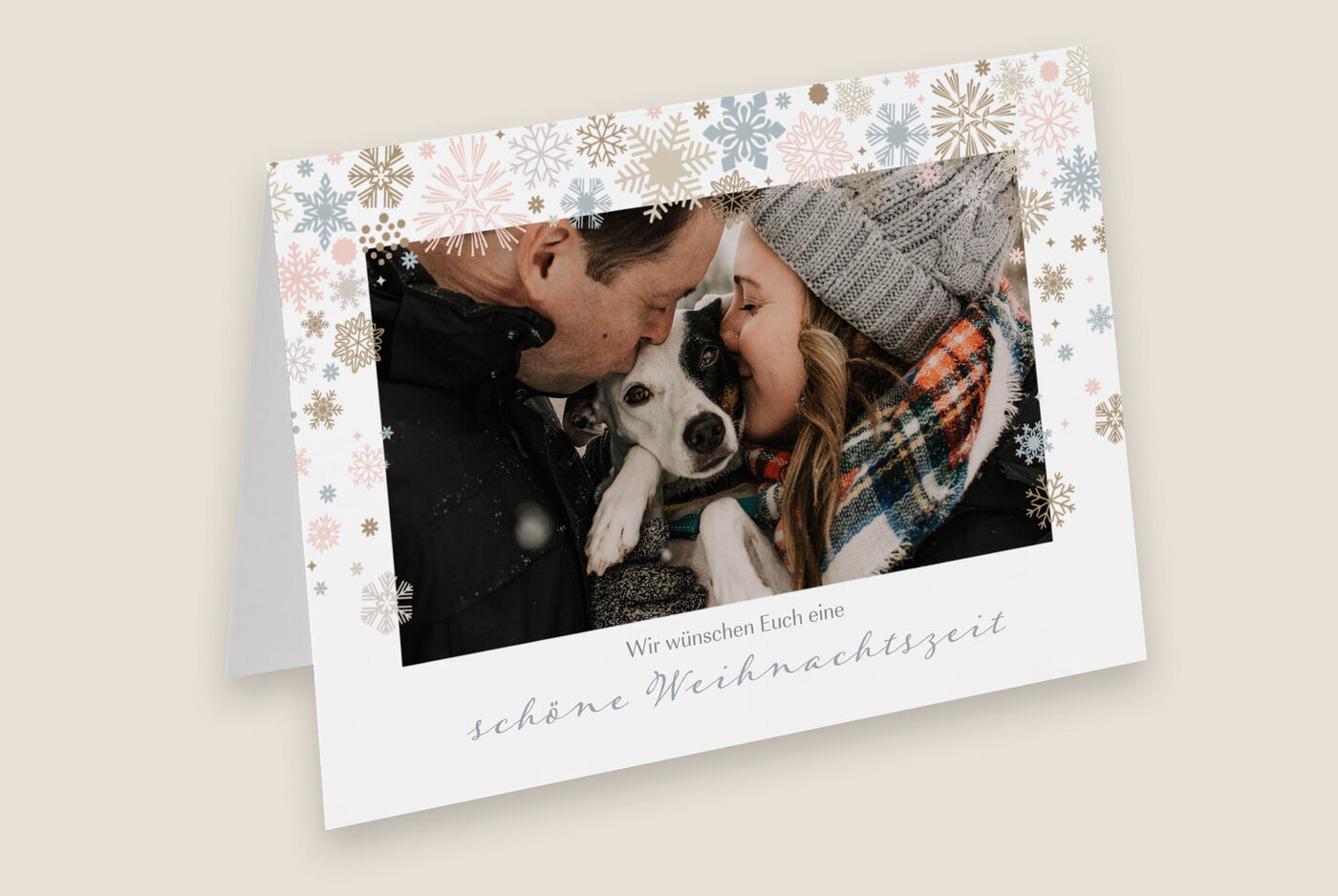 Eine liebevolle Grußkarte mit zwei Personen und einem Hund in winterlicher Kleidung umgeben von Schneeflockenmotiven.