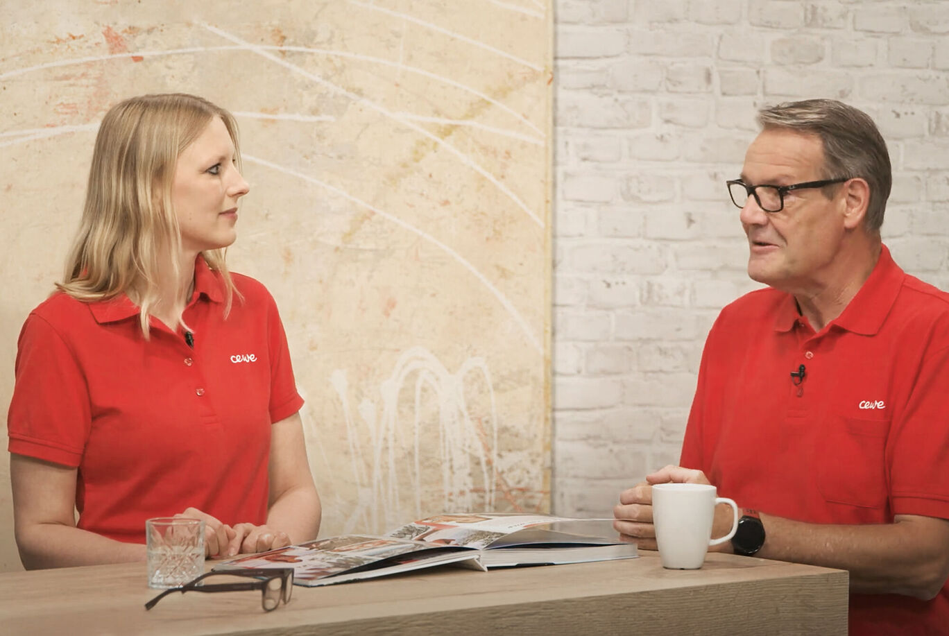 Zwei Personen in roten Poloshirts sitzen an einem Tisch und betrachten ein Fotobuch.