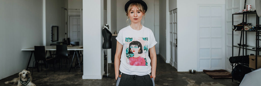 Junge Person steht in einem hellen Raum, trägt ein weißes T-Shirt mit buntem Grafikdruck und eine schwarze Mütze, im Hintergrund ein Hund und Möbel