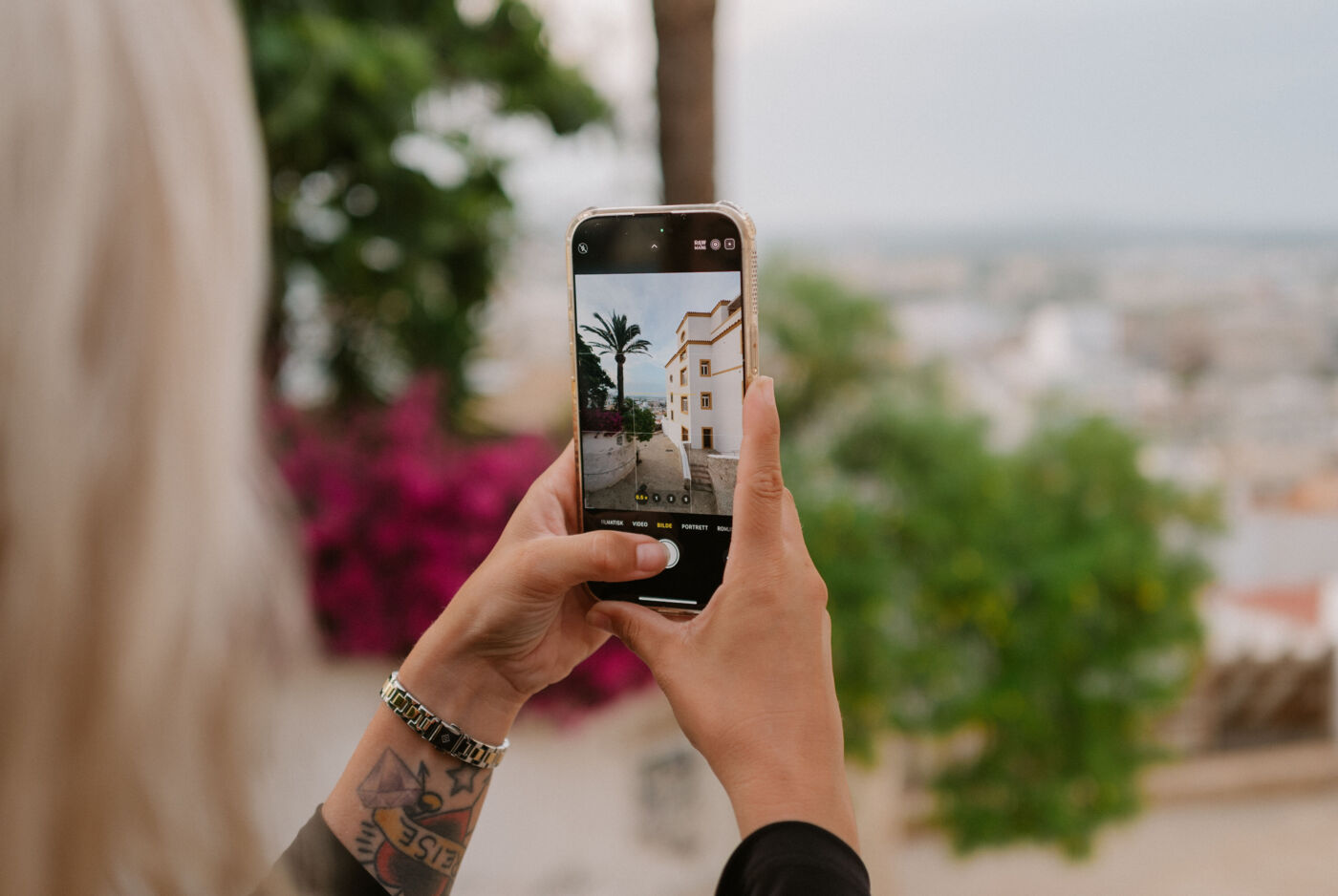 Frau hält Smartphone in Händen, um eine Gasse zu fotografieren. Sie ist gesäumt von einer Mauer, mit Palmen und Gewächsen, und einem Haus.