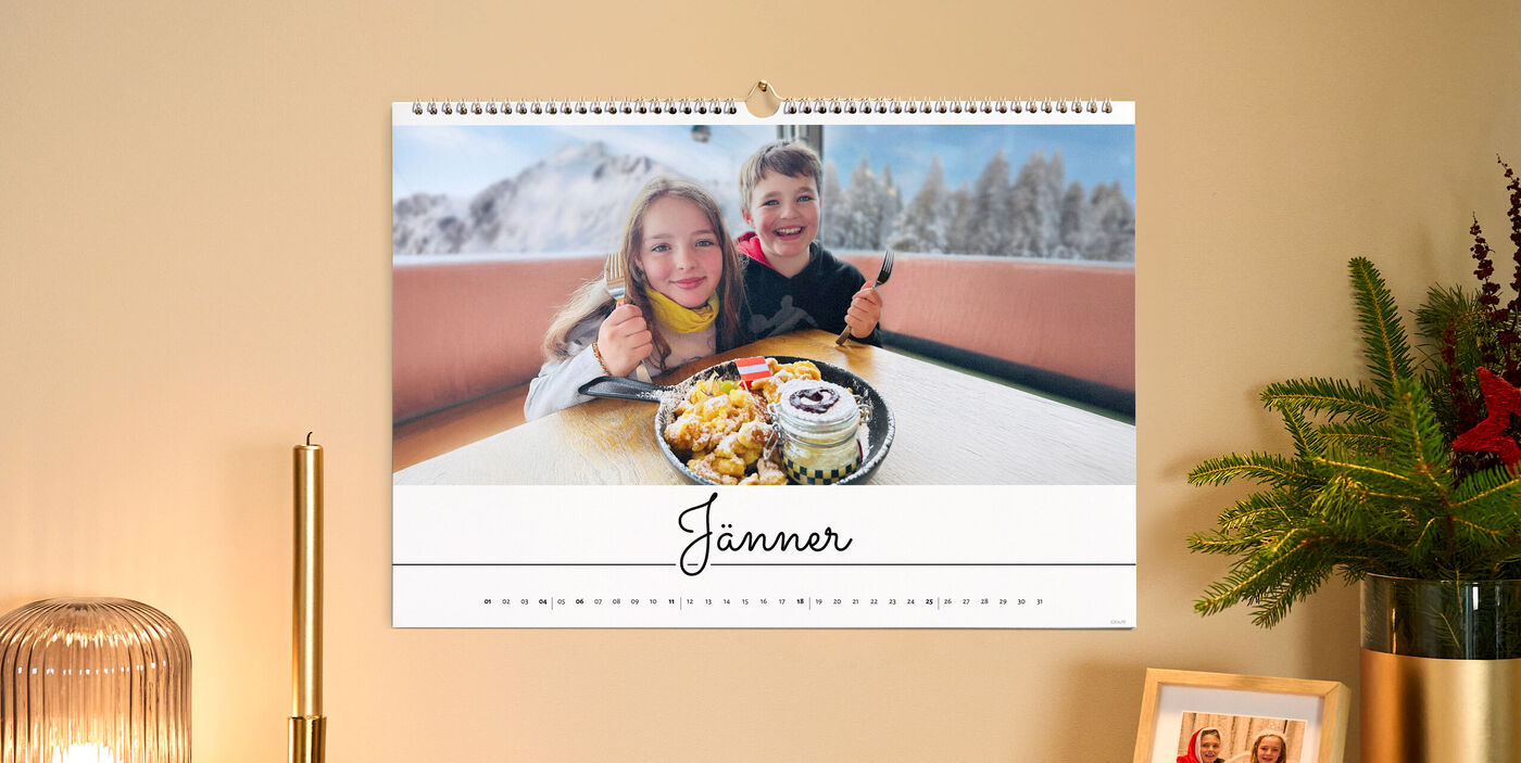 Ein Foto auf einem Kalender. Zwei Kinder sind lachend an einem Tisch vor einer Winterlandschaft im Hintergrund. Vor Ihnen ein Kaiserschmarrn