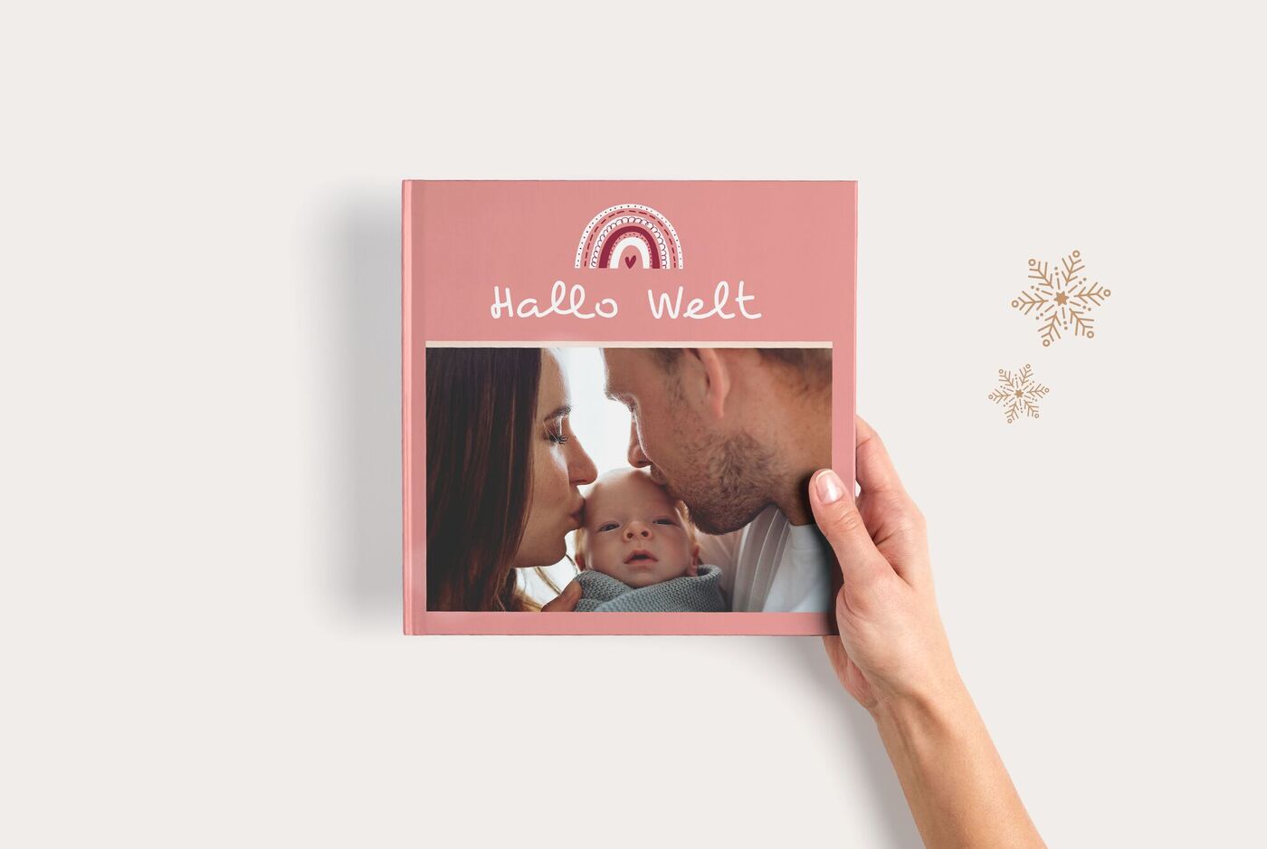Hand hält ein CEWE FOTOBUCH mit zwei Babys auf einer Decke und dem Titel Hallo Welt vor rosafarbenem Hintergrund