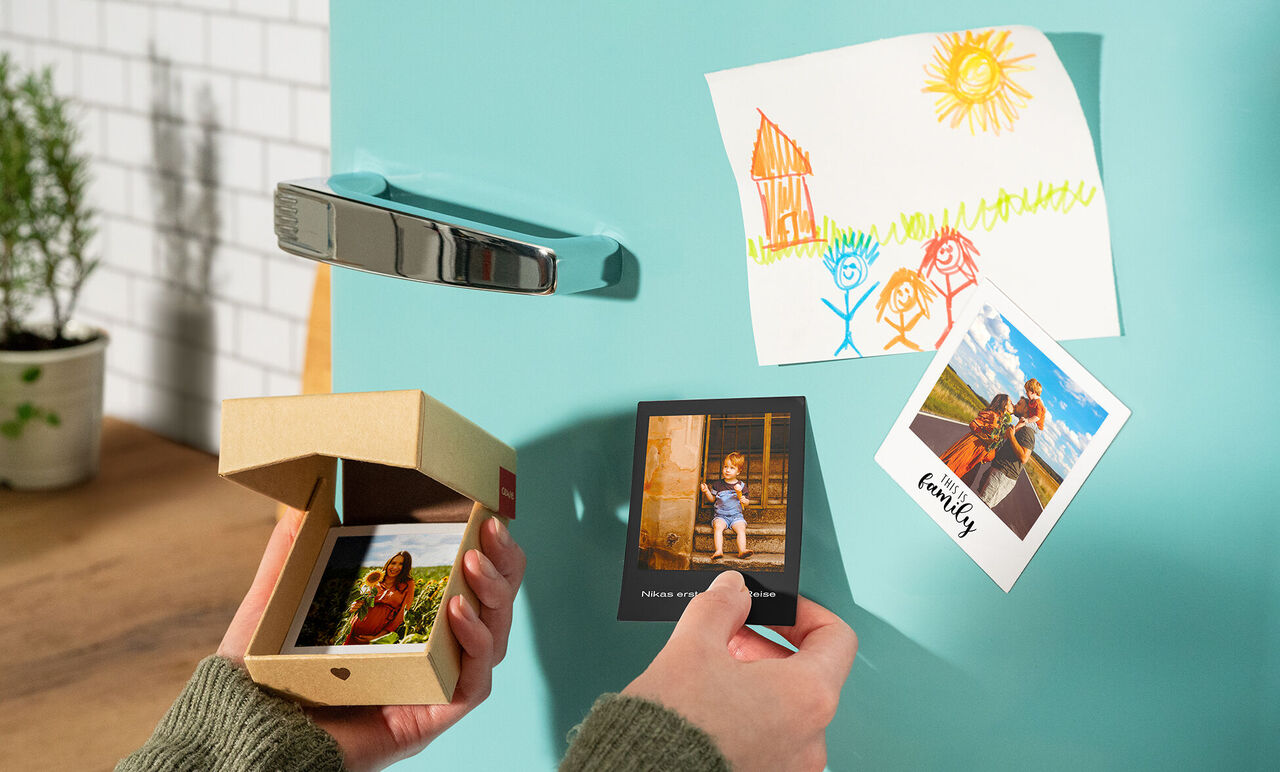 Eine Person hält eine kleine Box mit Fotos und die andere Hand zeigt ein Foto an einem Kühlschrank, dekoriert mit einem bunten Kinderbild.