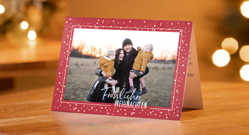 Aufgestellte Weihnachtsklappkarte auf einem Holztisch: Familienfoto mit zwei Erwachsenen, zwei Kindern im Freien, roter Rahmen mit Punkten