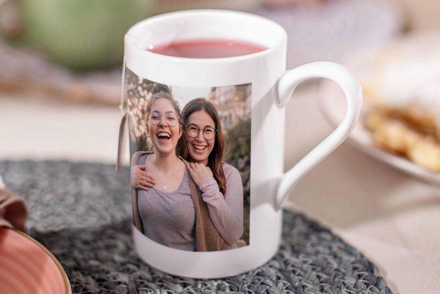 Eine personalisierte Tasse mit einem Foto von zwei Personen. Darauf ist eine warme, einladende Atmosphäre zu sehen. Die Tasse steht auf einem Tisch mit einem dekorativen Untersetzer und einer leichten Speise im Hintergrund. Eine schöne Möglichkeit, Lie