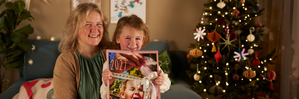 Ein Kind uns seine Mama stehen in einem Wohnzimmer neben einem geschmückten Weihnachtsbaum. Der Junge hält einen Fotokalender in die Kamera.