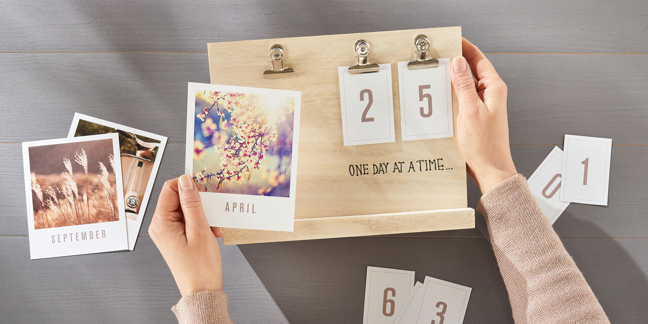 Zwei Hände befestigen einen Art Print mit Bildmotiv an einer Klemme des flexiblen Tischkalenders. Weitere Fotos und Ziffern liegen verteilt darum herum auf der Tischplatte.