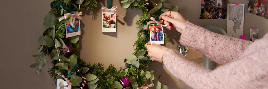 Eine Frau dekoriert mit Fotoanhängern einen weihnachtlich geschmückten Kranz, der an einer Wand hängt.