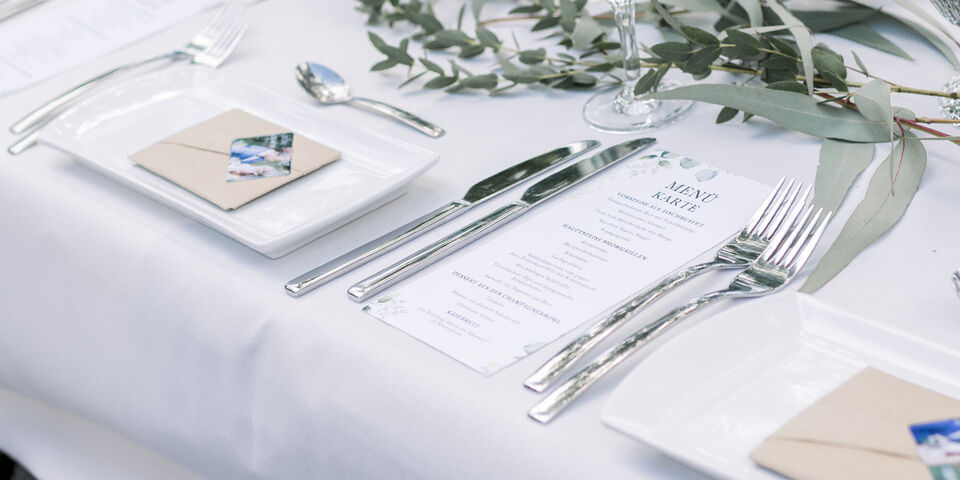 Elegantly arranged table setting with cutlery, plates, and menu card surrounded by greenery
