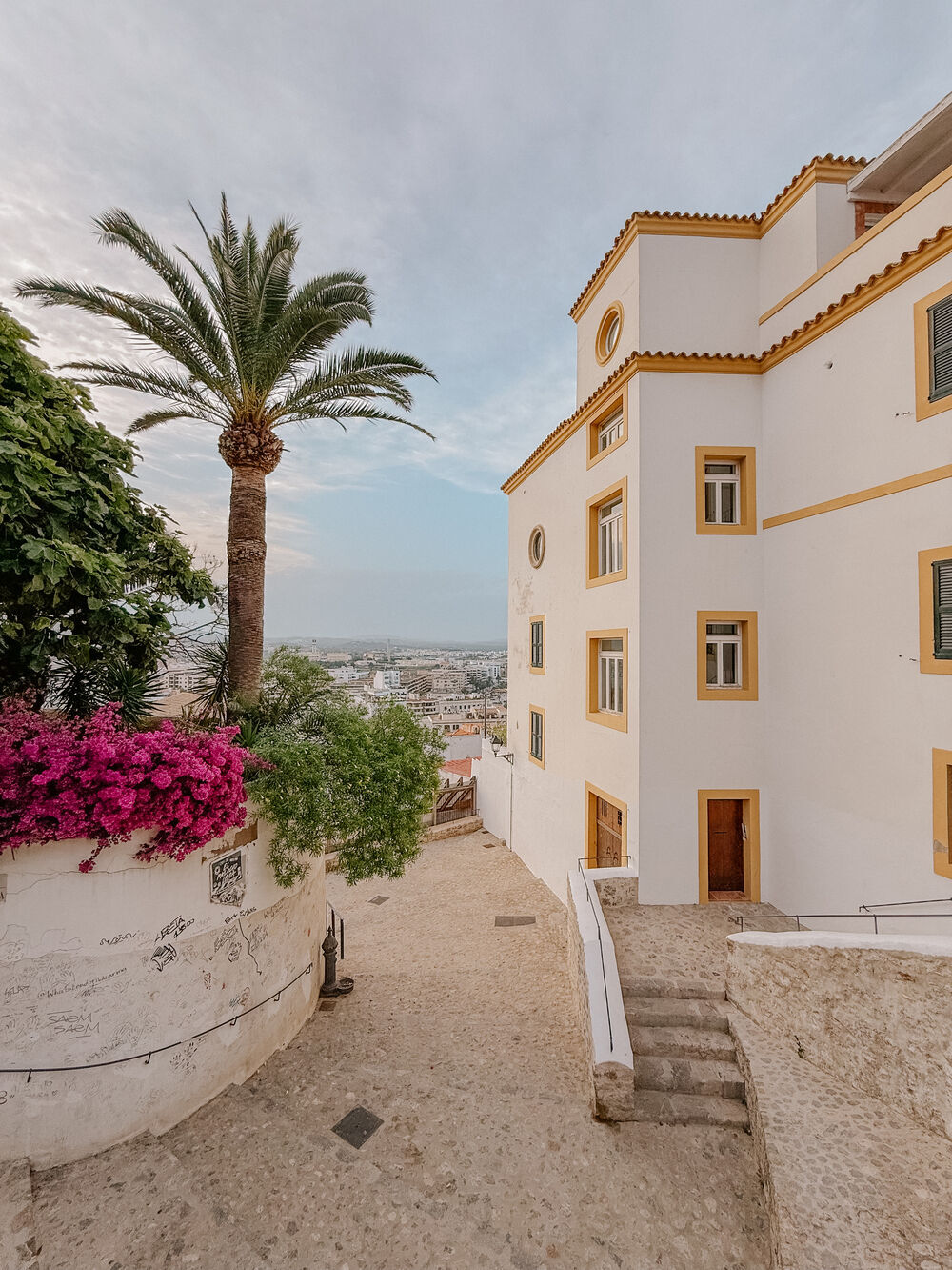 Helle, mediterrane Hausfassade mit gelben Fensterrahmen, Palme und blühenden pinken Blumen in einer Gasse mit Blick auf eine Stadt.