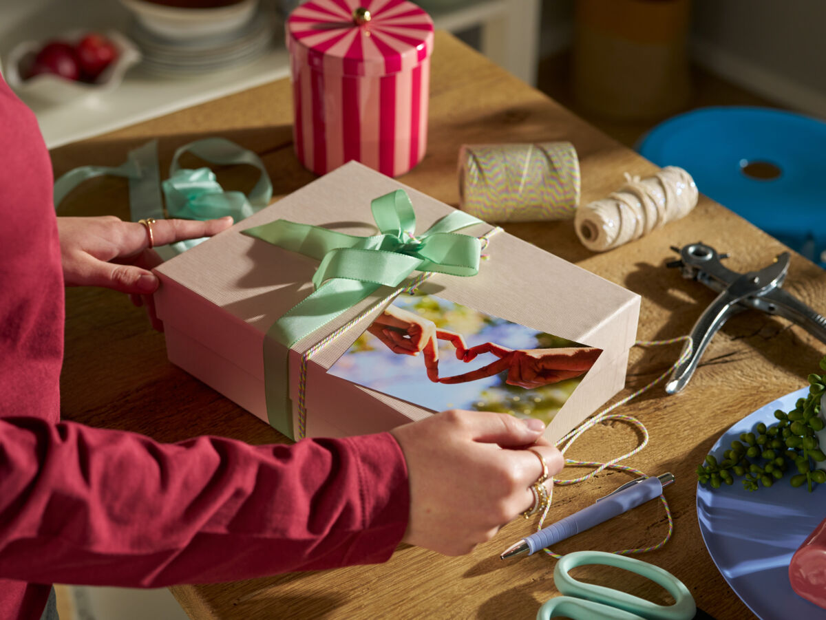 Auf einem Tisch liegt ein festlich eingepacktes Geschenk, das von einer Frau mit einer Fotopostkarte verziert wird.