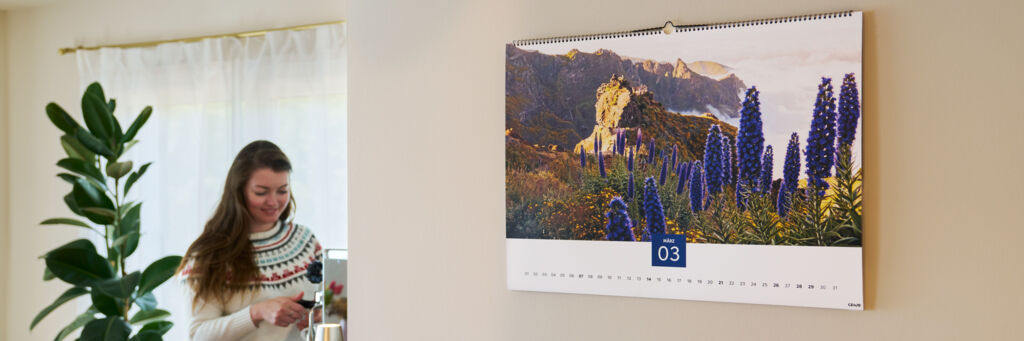 In einem Wohnzimmer hängt ein großer Wandkalender, im Hintergrund steht eine Frau an einer Kaffeemaschine und bereitet etwas zu.