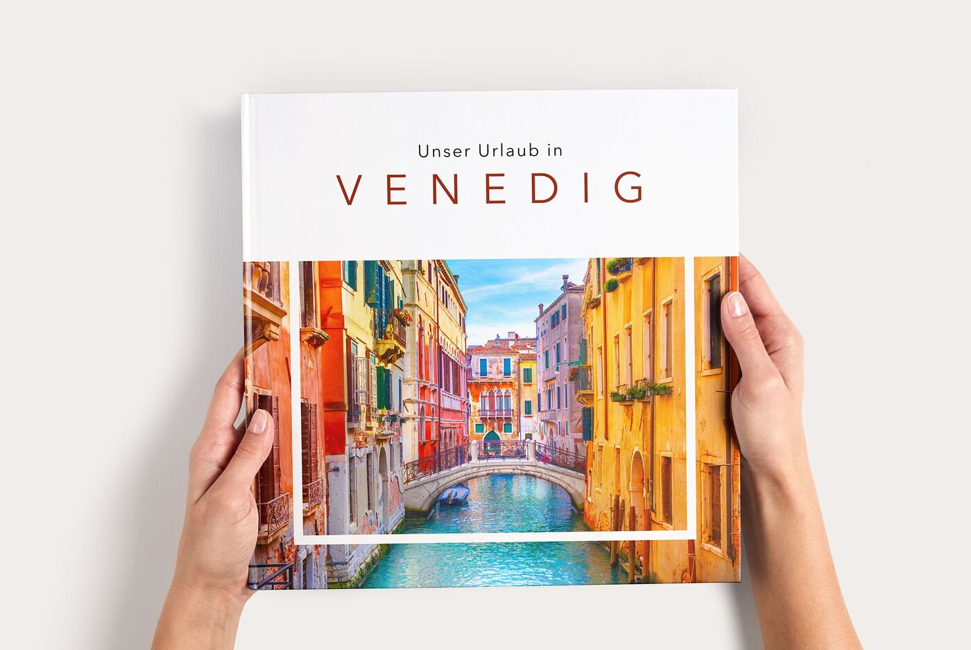 Zwei Hände halten ein CEWE FOTOBUCH mit dem Titel Unser Urlaub in Venedig und einem bunten Bild von Kanälen und Häusern in Venedig