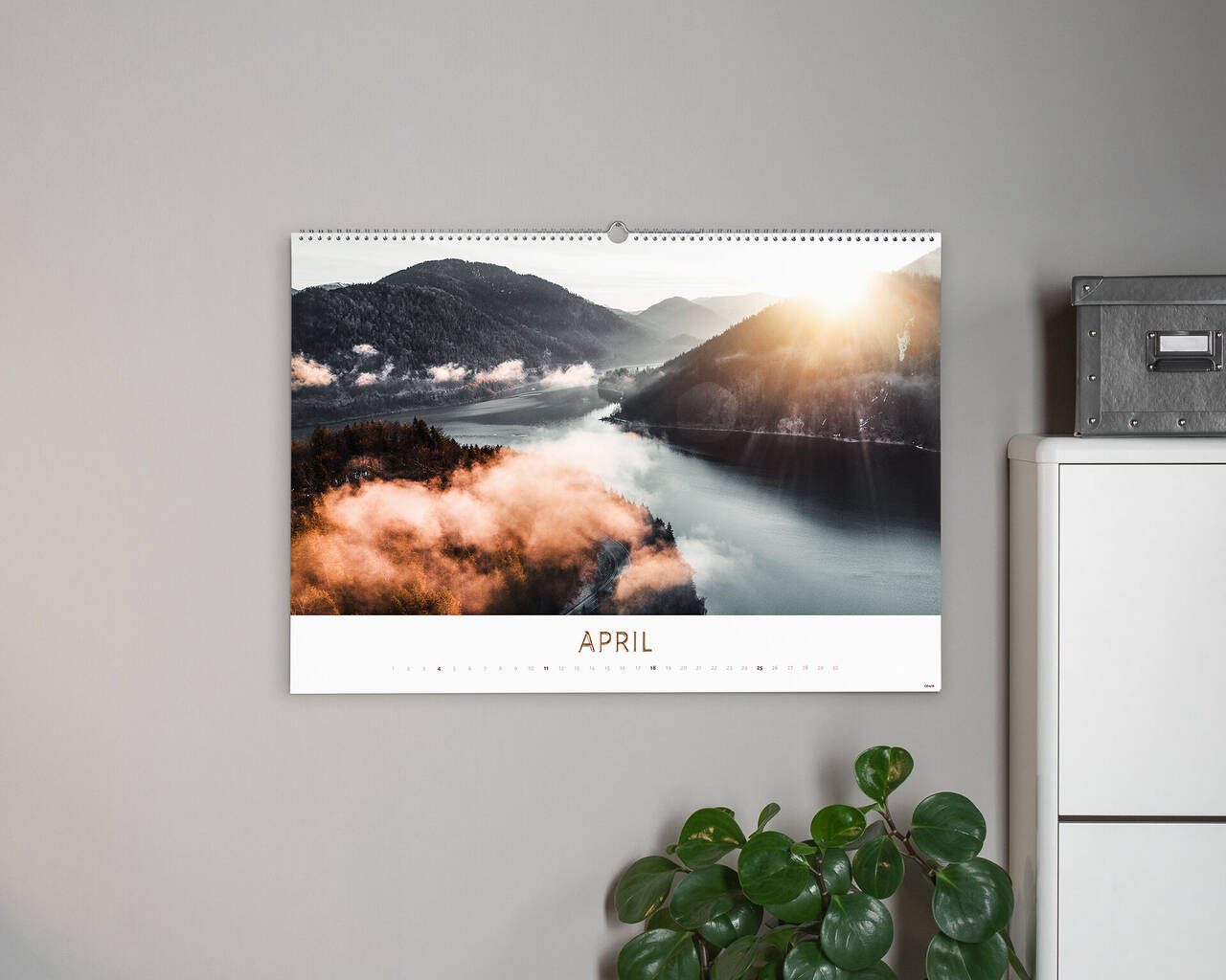 Wunderschöner Fotokalender mit einem landschaftlichen Bild für den Monat April. Der Kalender zeigt einen Sonnenaufgang über einem Nebel bedeckten See, umgeben von grünen Bergen. Ein kleiner Topf mit einer Pflanze steht daneben.
