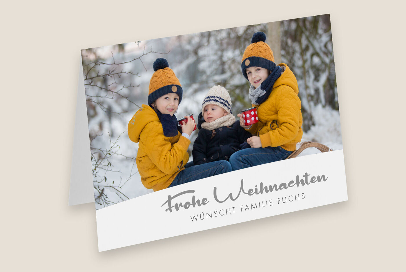 Drei Kinder in Winterkleidung mit Mützen sitzen im Schnee und halten Tassen. Sie wünschen frohe Weihnachten.