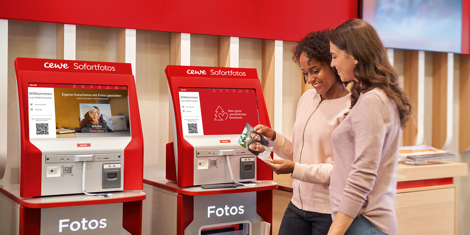 Zwei Personen stehen an einem CEWE Sofortfoto-Automaten und halten Fotos in der Hand