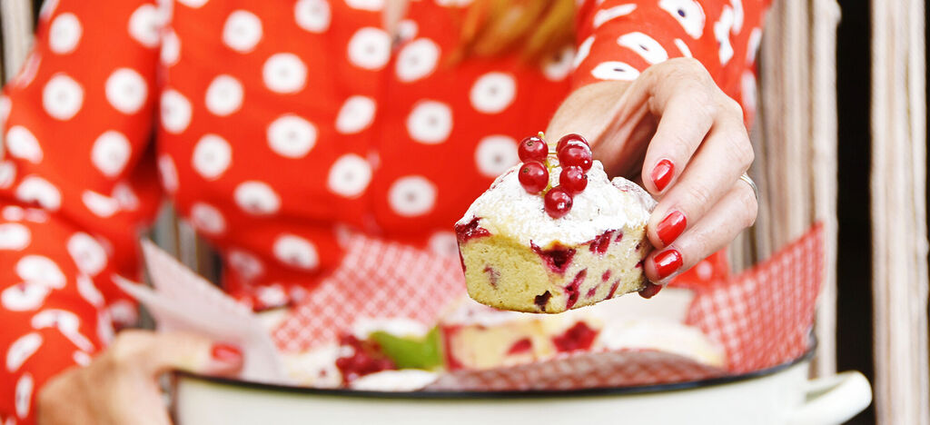 Eine Person mit rotem gepunktetem Shirt hält ein Stück Kuchen mit Johannisbeeren und Puderzucker in der Hand. Der Hintergrund ist hell und harmonisch.