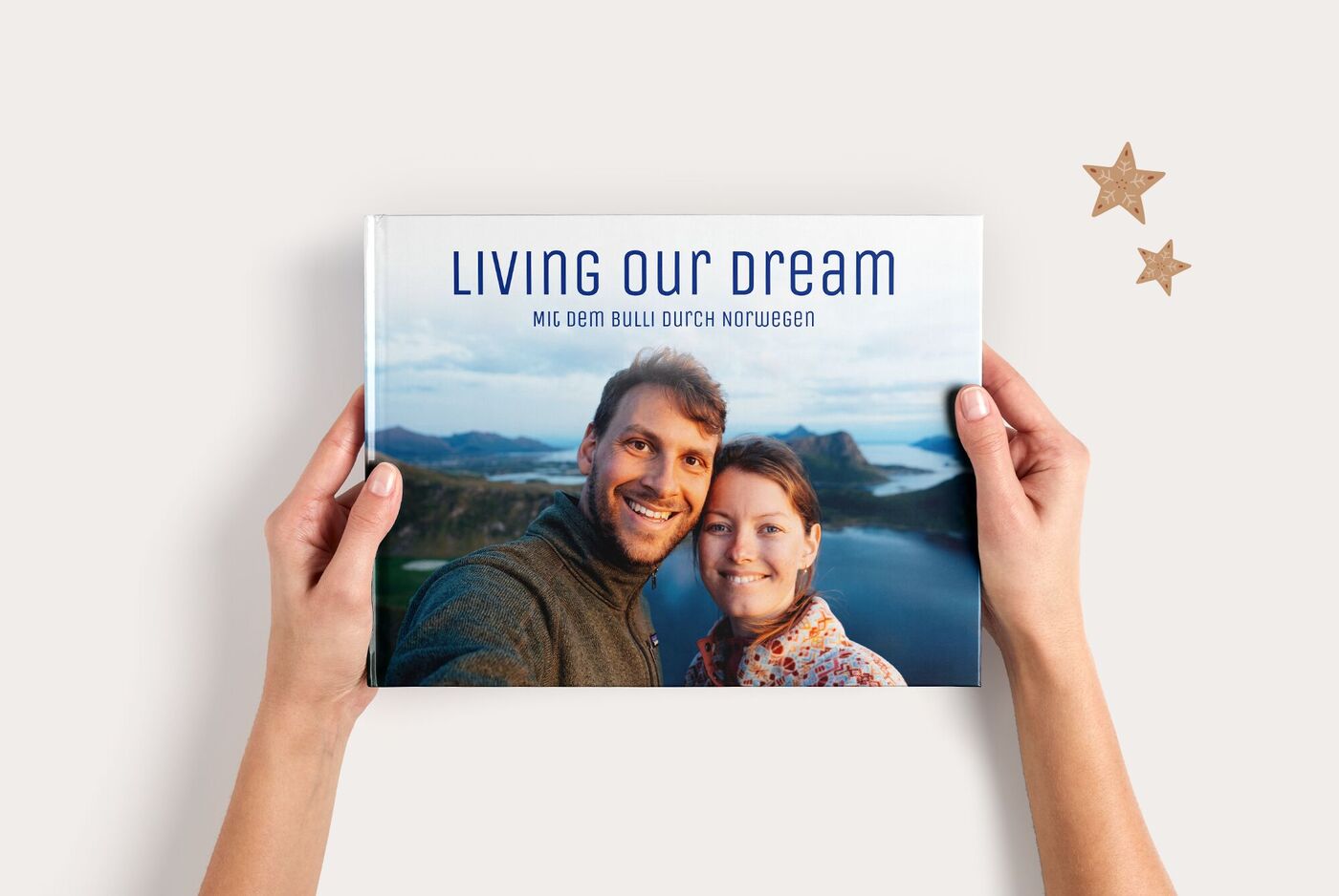 Zwei Hände halten ein CEWE FOTOBUCH mit dem Titel Living our dream und Bullireise durch Norwegen, im Hintergrund Berge und Wasser