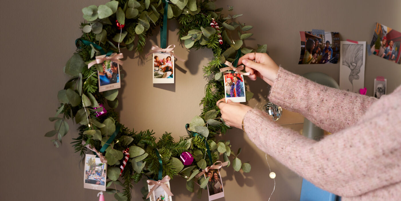 Eine Frau dekoriert mit Fotoanhängern einen weihnachtlich geschmückten Kranz, der an einer Wand hängt.