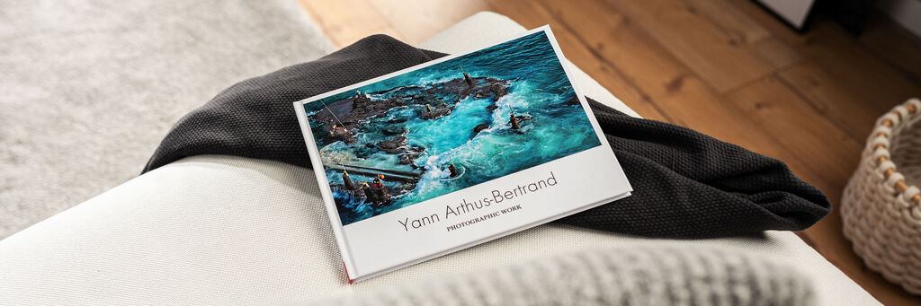 Ein CEWE Fotobuch auf einer Couch, offen mit Bildern von einem wunderschönen Strandurlaub vor einer sanften Wellenlandschaft.