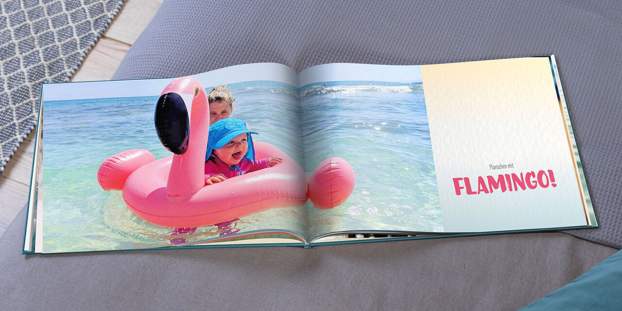 Ein aufgeschlagenes CEWE FOTOBUCH zeigt Hannah auf einem aufblasbaren Flamingo im Pool. Auf der rechten Seite des Buchs steht "Planschen mit FLAMINGO!".