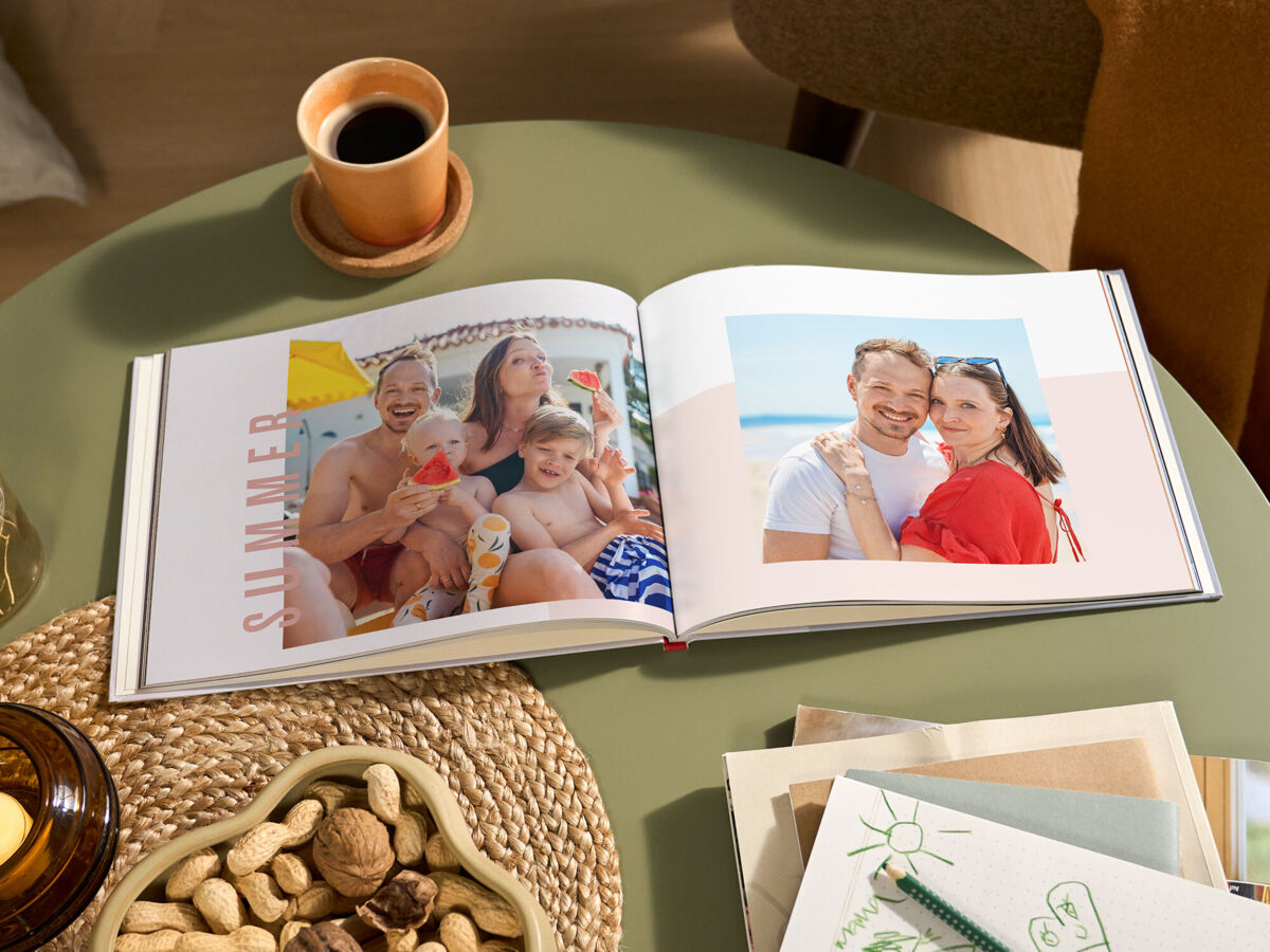 Aufgeschlagenes Fotoalbum mit sommerlichen Familienfotos und der Aufschrift „SUMMER“ auf einem grünen Tisch; daneben stehen eine Tasse Kaffe