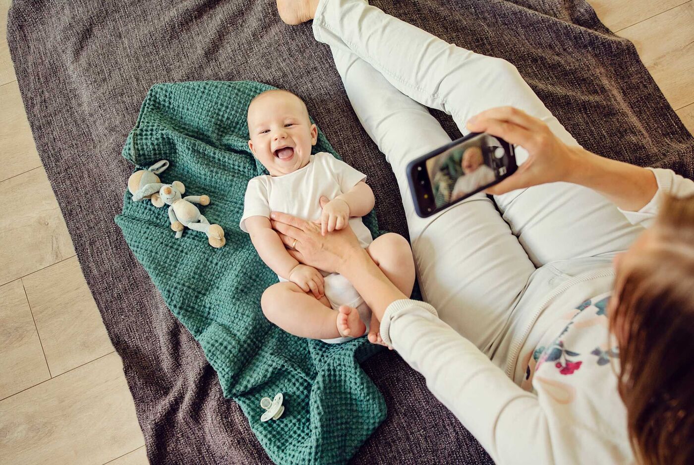 Eine liebevolle Momentaufnahme einer Mutter, die auf dem Boden sitzt und ein Baby lächelnd in die Kamera ihres Handys anlächelt. Im Hintergrund liegen ein grünes Tuch und ein paar Spielzeuge.