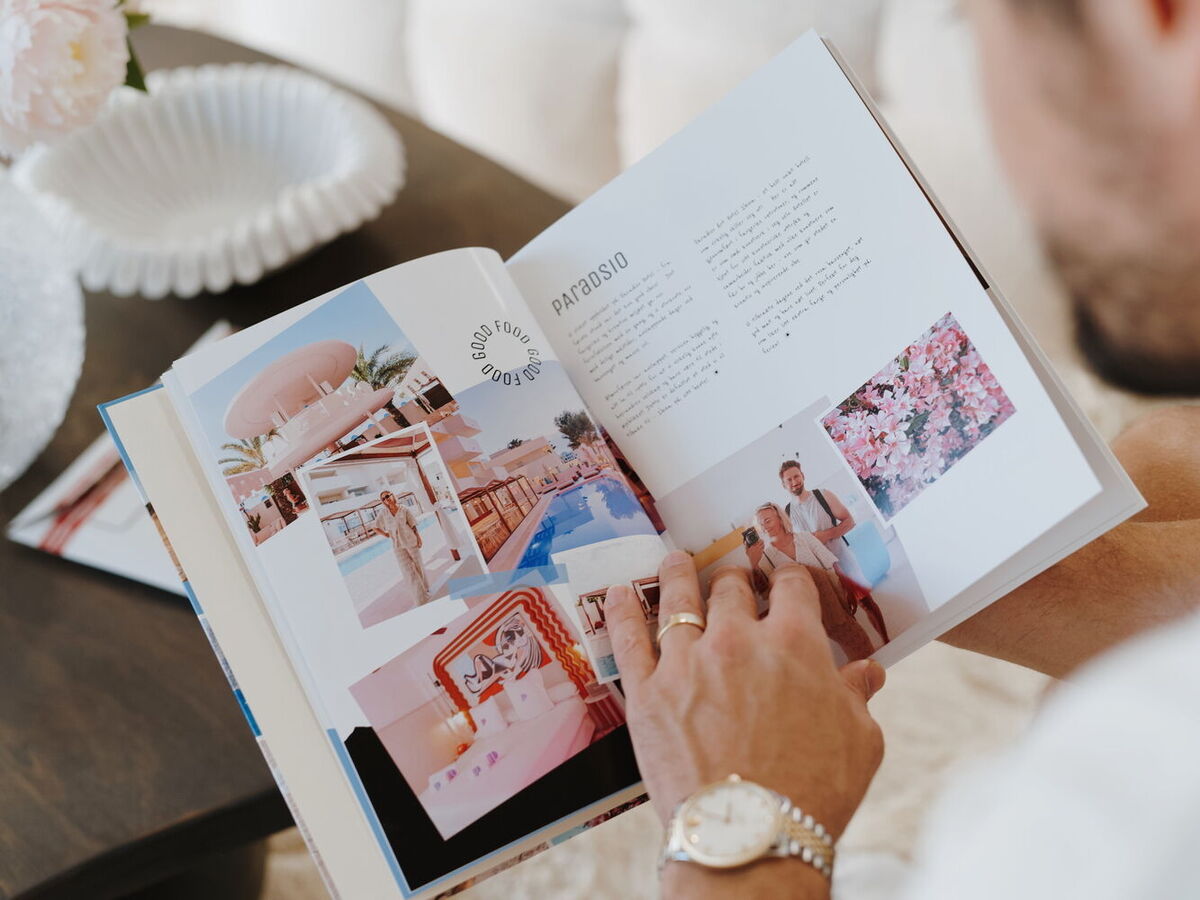 Person blättert in einem Fotobuch mit Urlaubsbildern und Texten. Im Hintergrund ist unscharf ein Tisch zu erkennen.