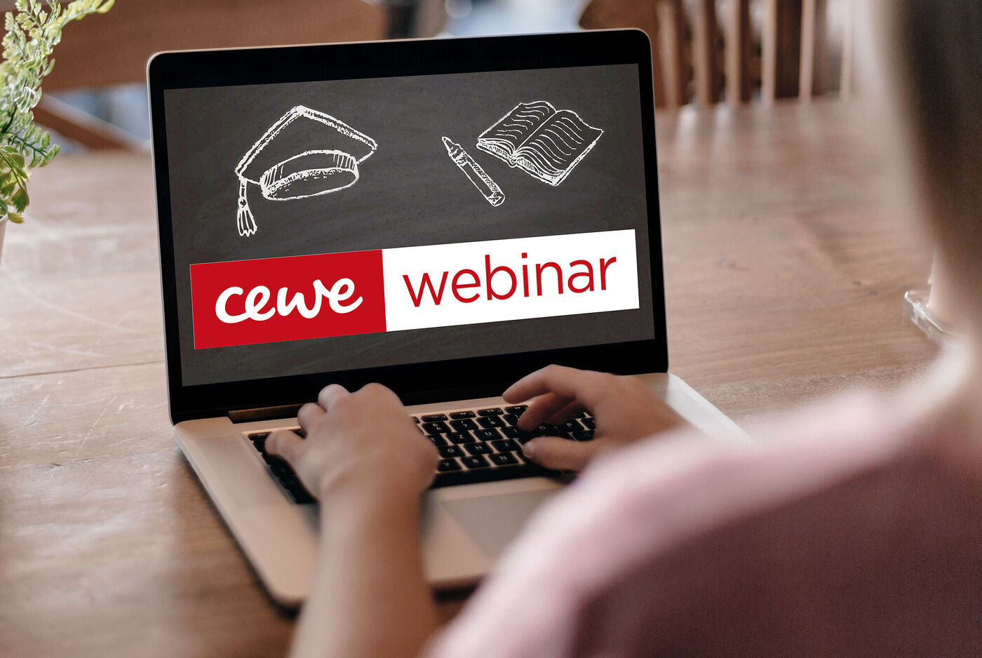 Person, die an einem Laptop sitzt und an einem CEWE Webinar teilnimmt, Schreibtisch mit Notizen und Zeichenmaterial.