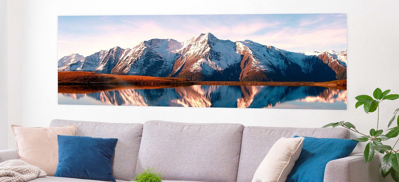 Panoramaaufnahme eines majestätischen Bergpanoramas mit schneebedeckten Gipfeln, die sich im ruhigen Wasser eines Sees spiegeln. Ein warmes Licht erleuchtet die Landschaft.